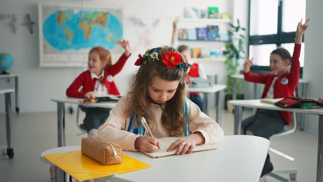 Little sad Ukrainian girl writing in classroom during class, concept of enrolling Ukrainian kids to schools.