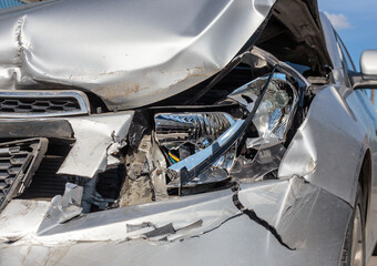 The front part of the silver car is damaged as a result of an accident on the road. The front part of the car is silver, damaged and broken as a result of an accident. Hood, headlight, bumper. 