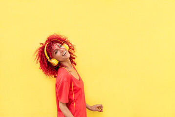 happy latin woman with afro red hair listening to music with yellow headphones over yellow background wearing red shirt looking at the camera