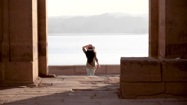 Caucasian woman with hat running through the ruins of Temple of Philae with lake in background in Aswan, Egypt.