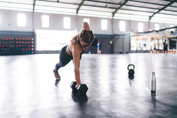 Fototapeta na wymiar Strong Caucaisan athlete in sportswear doing pushup plank at one hand holding dumbbells in gym studio, fit girl in tracksuit using hard equipment for training body muscles during intensive crossfit