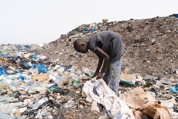 Young African garbage collector filling a plastic bag with reusable objects in an urban landfill;...