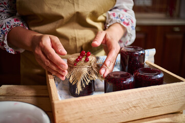 Woman hands next to a jar with jam, ornate with red currant berries on the burlap on the cover