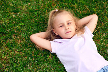 Little blonde girl lies on the grass in a summer city park and rests looking at the camera