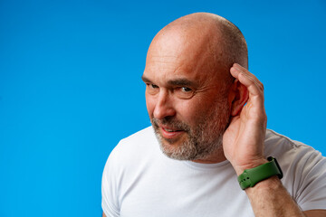 Handsome middle age man holding hand near ear trying to listen against blue background