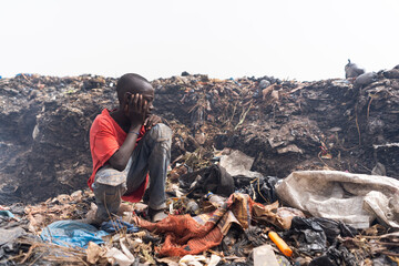 Poor African slum boy sitting in front of a large pile of garbage in the middle of a huge urban...