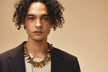 Serious stylish awesome tanned curly man in classic brown jacket ethnic necklace posing isolated on over beige pastel background. Fashion New Collection offer. Retro style concept. Free place for ad
