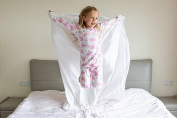 Beautiful little girl in pajamas jumping on the bed with white blanket at home. Stay at home during...