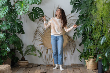 Plant lover. Happy young German woman in cozy tropical indoor garden, taking care of monstera plant, touching lush green leaves and smiling while standing in greenhouse. Hobby, gardening concept © DimaBerlin