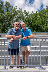 Two Caucasian men, a senior gay married couple, lean against a railing in a stadium and are looking off to the distance.