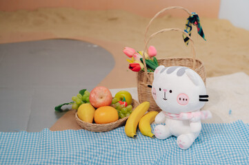 Obraz premium Cat doll with roses in a basket and group of fruit on blue fabric and sand background.