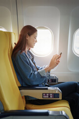 Attractive portrait of Asian woman sitting at window seat in economy class using mobile phone...