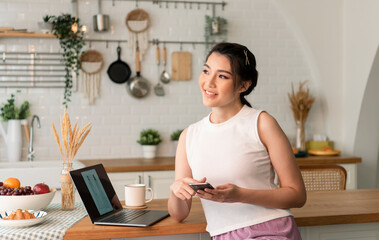 Smiling young asian woman using mobile phone while sitting in kitchen room at home with laptop computer.