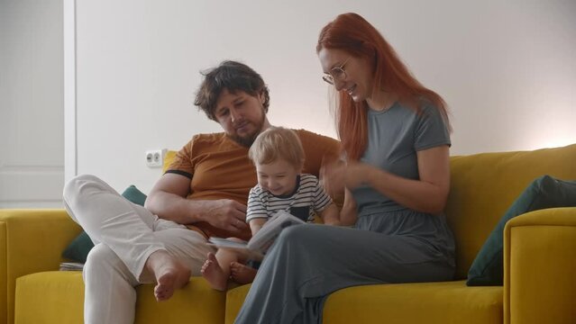 Family Sitting On Yellow Couch And Teaching The Baby How To Read - The Baby Drawing In A Book