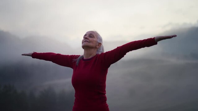Senior woman doing breathing exercise in nature on early morning with fog and mountains in background.