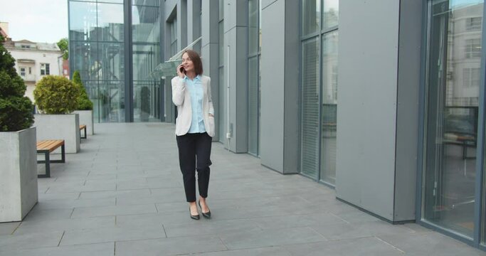 Young pretty female talking on mobile phone and walking the street at office center. Beautiful busineswoman having cellphone call ad walk to work or at business meeting. Calling on telephone.