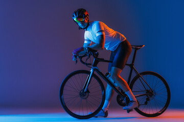 One male cyclist riding bicycle wearing cycling shorts and protective helmet isolated on dark blue background in neon. Concept of sport, speed, energy