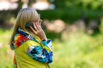 brightly dressed blonde mature woman talking on smartphone in garden, online communication concept