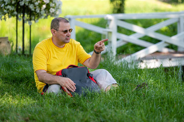 Naklejka na ściany i meble a senior man with a backpack sits on the grass in a park on a sunny day