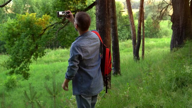 Male tourist with backpack while traveling takes pictures with smartphone summer landscape in forest. Travel blogger shoot video, make photos