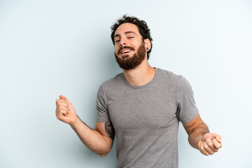 young adult bearded man smiling, feeling carefree, relaxed and happy, dancing and listening to...