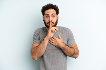 young adult bearded man looking serious and cross with finger pressed to lips demanding silence or...
