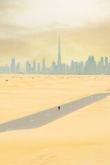 View from above, stunning aerial view of a person walking on a deserted road covered by sand dunes with the Dubai Skyline in the distance. Dubai, United Arab Emirates.