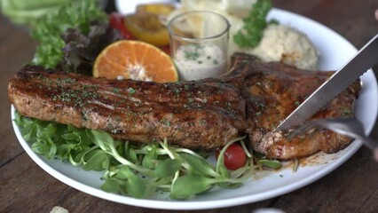 Cutting Pork t-bone with knife and fork meat fat and bone steak with salad and mashed potato sides