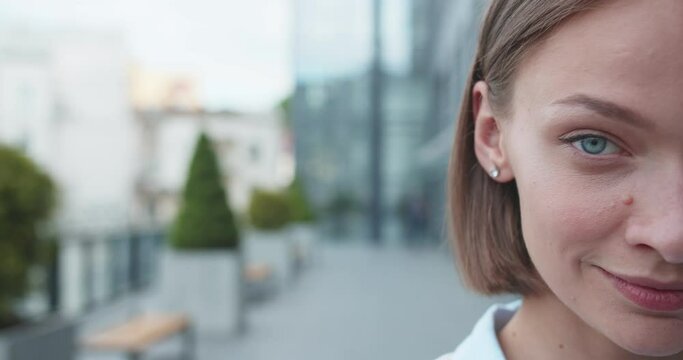 Close up of half face of young Caucasian beautiful woman looking straight to camera and smiling while standing outdoor in modern city street. Portrait of pretty female in business style.