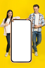 Pointing at white screen caucasian man and asian woman standing leaned on huge smartphone with...