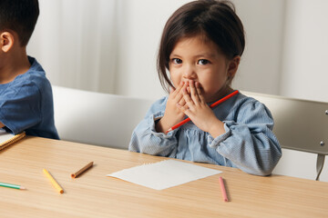 a cute, pleasant little girl of pre-school age sits at a table and draws with colored pencils, smiling covering her face with her hands. Themes of hobby, development and training - obrazy, fototapety, plakaty