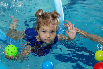 Little curly toddler girl learns to swim in swimming pool using foam stick.