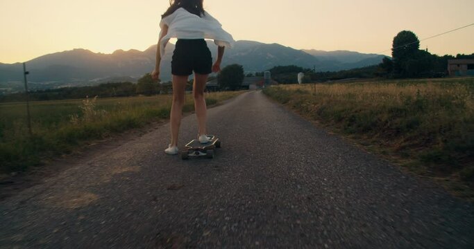 Young carefree and stylish adult woman ride on skate longboard at sunset along meadow. Trendy teenager girl skateboarding with mountain view. Concept of youth and freedom on countryside