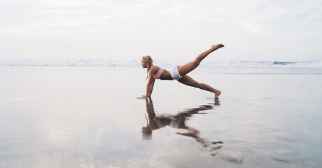 Full length view, Caucasian female with sporty body doing stretching exercises on seashore while...