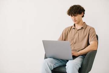 Attractive young man using laptop while sitting in armchair against white wall, copy space. Millennial male communicating online, working or studying remotely on portable pc