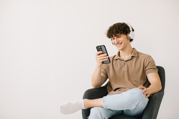 Attractive young man using laptop, wearing headphones while sitting in armchair against white wall, copy space. Millennial male communicating online or relaxing