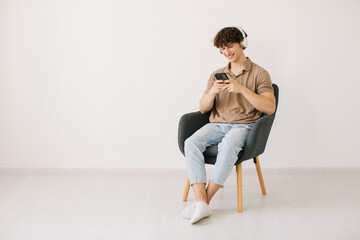 Obraz premium Attractive young man using laptop, wearing headphones while sitting in armchair against white wall, copy space. Millennial male communicating online or relaxing