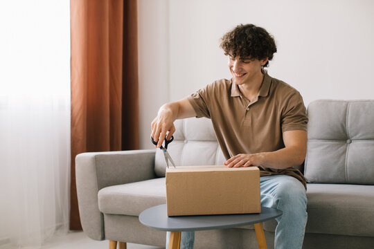 Happy young smiling curly man opening box with ordered goods gifts, presents at home on couch. Online shopper male customer opening online shop parcel.