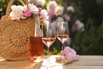 Bottle and glasses of rose wine near beautiful peonies on wooden table in garden