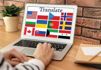 Translator using modern laptop with images of different flags on screen at table indoors, closeup
