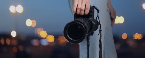Professional photographer with modern camera and blurred view of beautiful city in evening. Banner design - obrazy, fototapety, plakaty