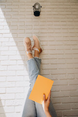 Woman holding a yellow notebook with her legs up in blue jeans and pink sneakers against a brick wall background
