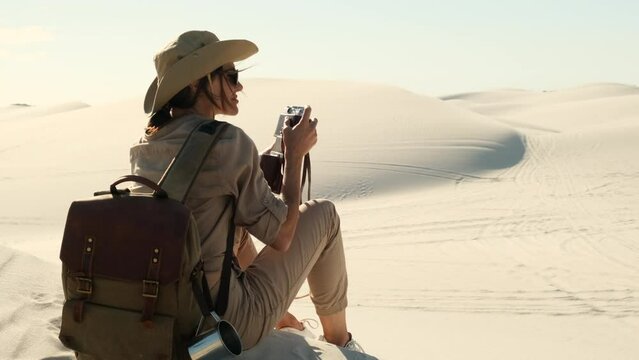 woman in travel clothes sits on sand dune in the desert and takes a photo on a film camera. travel female enjoys traveling alone through the desert in africa. girl in boots and a hat sits on the sand
