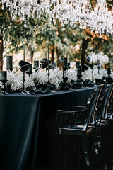 Serving and decoration of the banquet table in black with white flowers, black candles, and crockery. Crystal chandeliers illuminate the table from above.