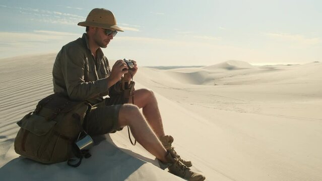 man in travel clothes sits on a sand dune in the desert and takes a photo on a film camera. travel man enjoys traveling alone through the desert in africa. a man in boots and a hat sits on the sand