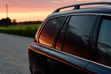 SUV on off-road rocky road. A beautiful red summer evening sunset is reflected in the SUV's tinted...