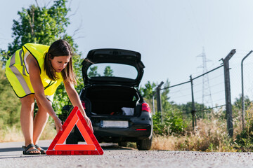 Woman with yellow reflective vest placing the emergency warning system for her broken down car. Young girl supporting a warning triangle for broken down vehicles.