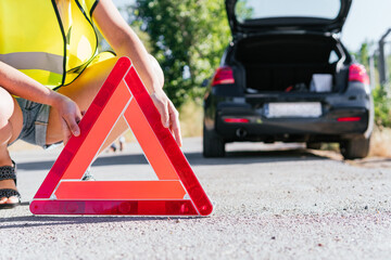 Woman with yellow reflective vest placing the emergency warning system for her broken down car. Close up image of a young girl supporting a warning triangle for broken down vehicles.