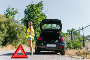 Woman in yellow reflective vest standing desperate with her broken car on the shoulder. Young girl worried about her broken car while waiting for insurance assistance with an emergency triangle on.