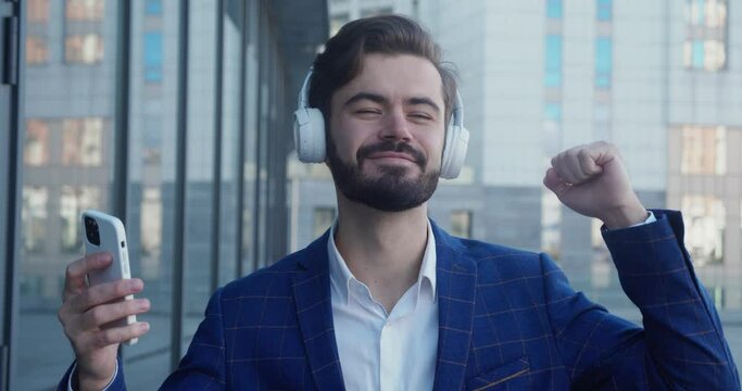 Caucasian happy beautiful man in suit in headphones dansing on street and listening to music outdoors. Joyful male in good mood enjoying songs in town. Close up portrait. Leisure concept.